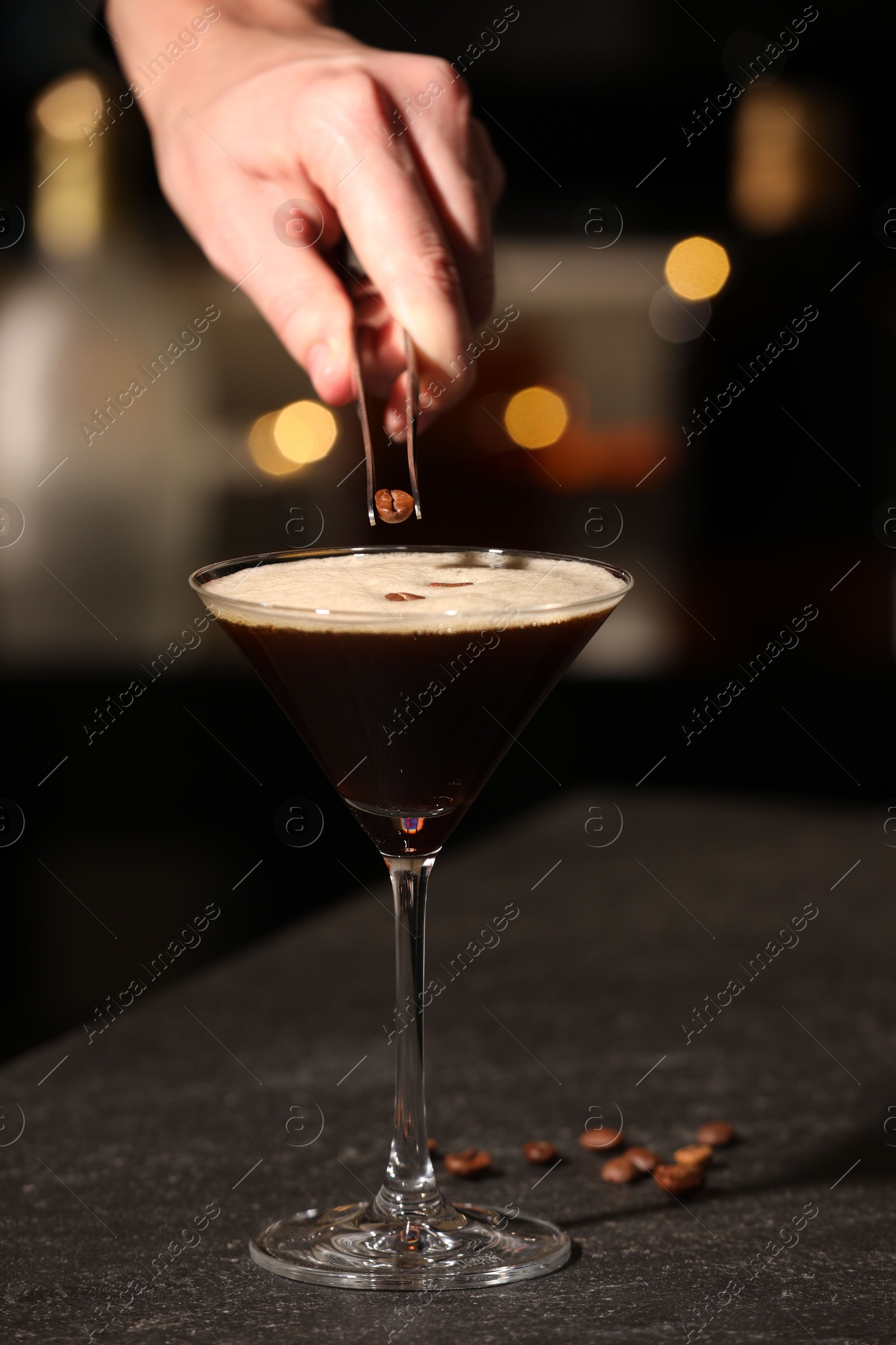 Photo of Bartender putting coffee bean into glass with delicious espresso martini at dark table against blurred lights, closeup