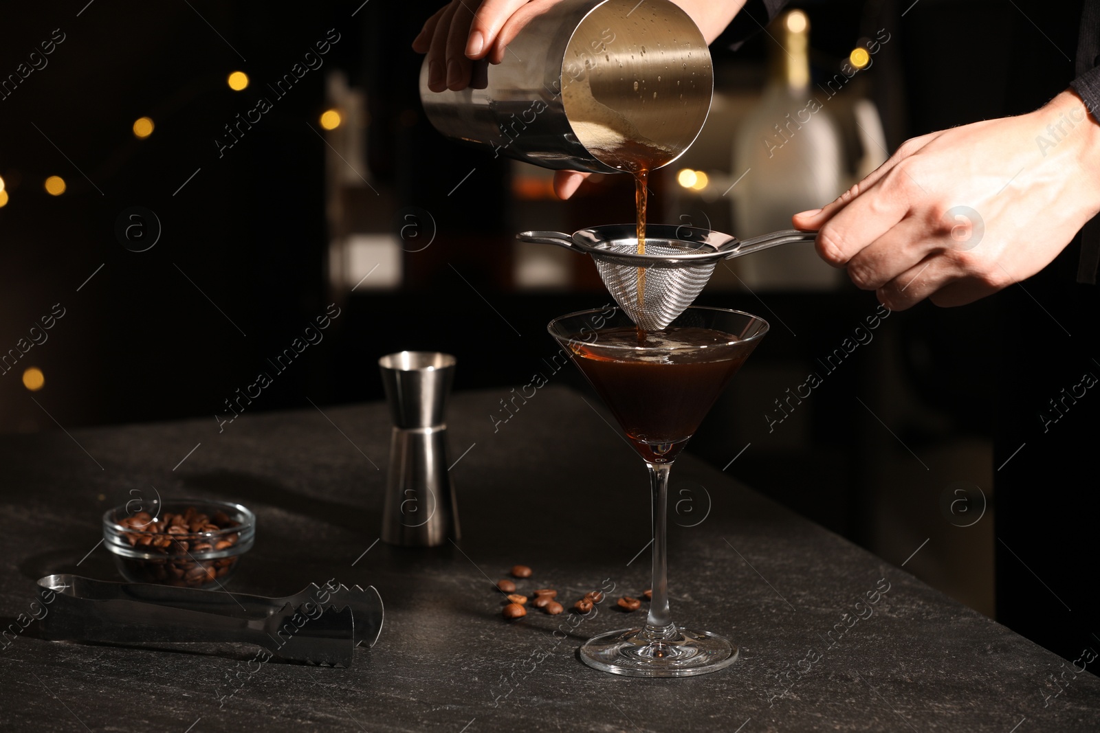 Photo of Bartender making delicious espresso martini at dark table against blurred lights, closeup