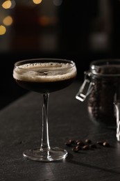 Photo of Glass with delicious espresso martini and coffee beans on dark table against blurred lights