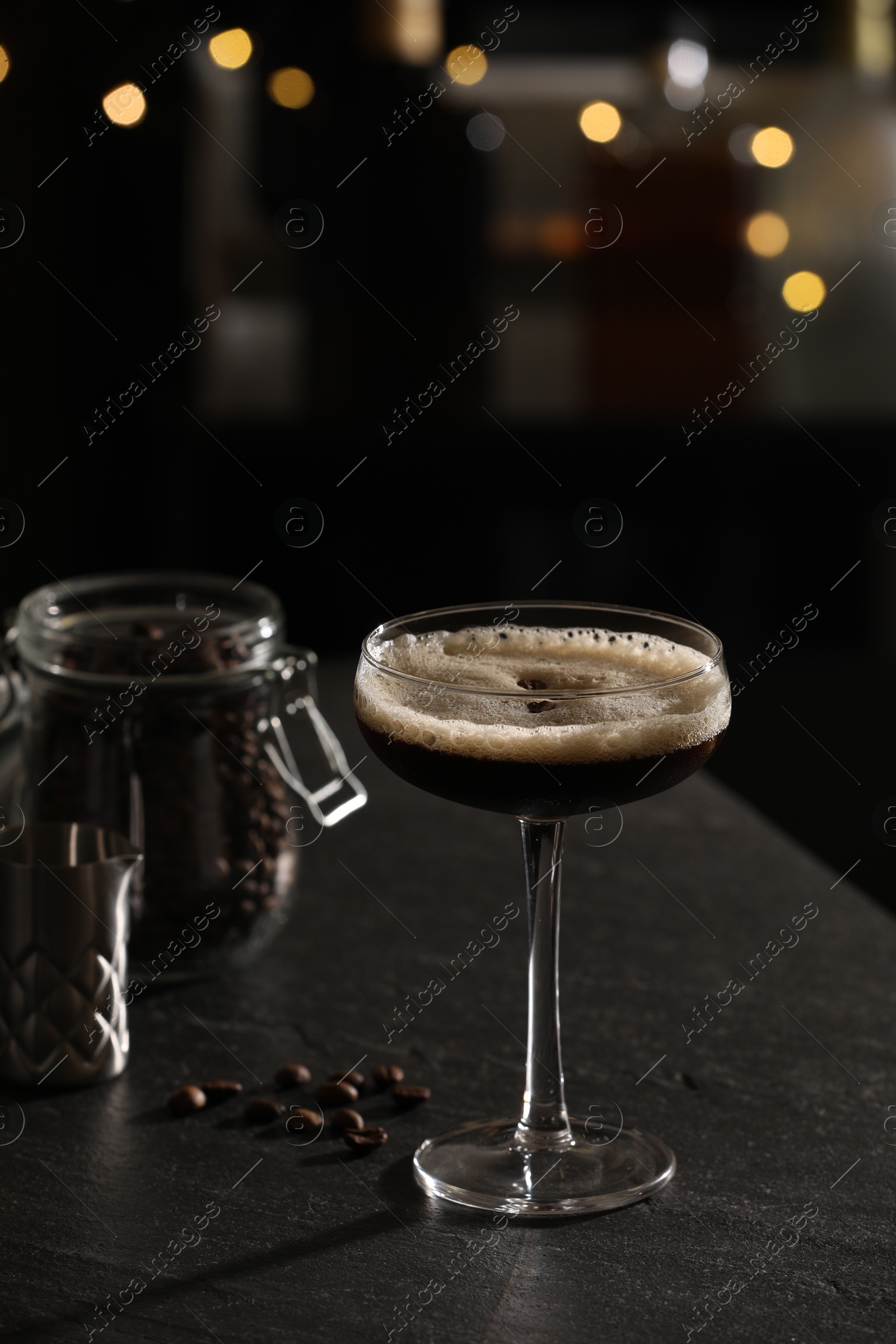 Photo of Glass with delicious espresso martini and coffee beans on dark table against blurred lights