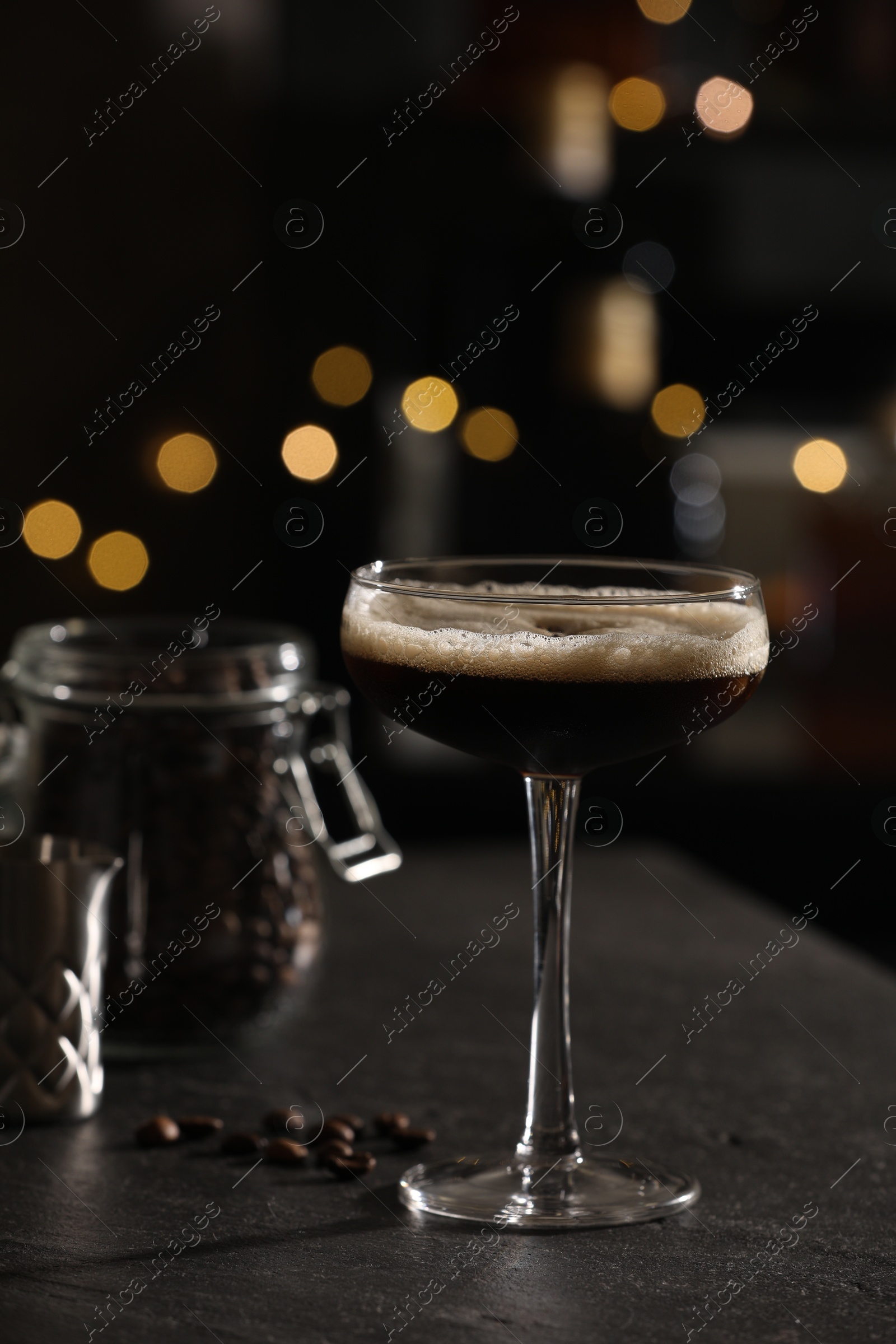 Photo of Glass with delicious espresso martini and coffee beans on dark table against blurred lights