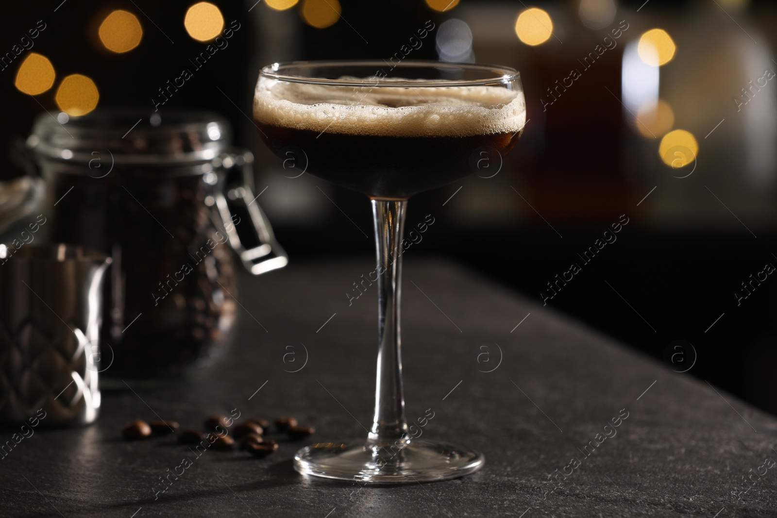 Photo of Glass with delicious espresso martini and coffee beans on dark table against blurred lights, closeup