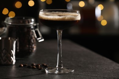 Glass with delicious espresso martini and coffee beans on dark table against blurred lights, closeup