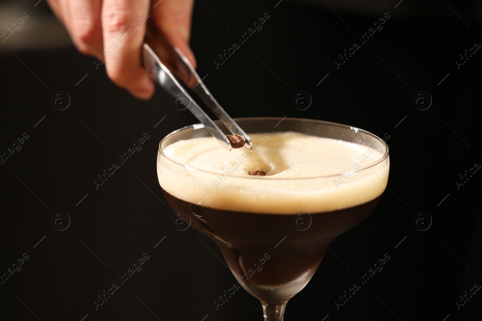 Photo of Bartender putting coffee bean into glass with delicious espresso martini on black background, closeup