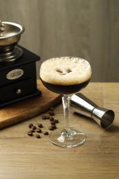 Photo of Delicious espresso martini in glass, coffee beans, manual grinder and jigger on wooden table