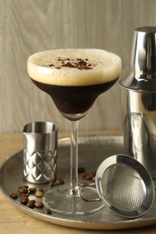 Photo of Tray with delicious espresso martini in glass, sieve, shaker and coffee beans on wooden table, closeup