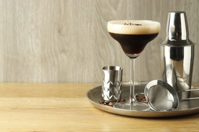 Tray with delicious espresso martini in glass, sieve, shaker and coffee beans on wooden table. Space for text