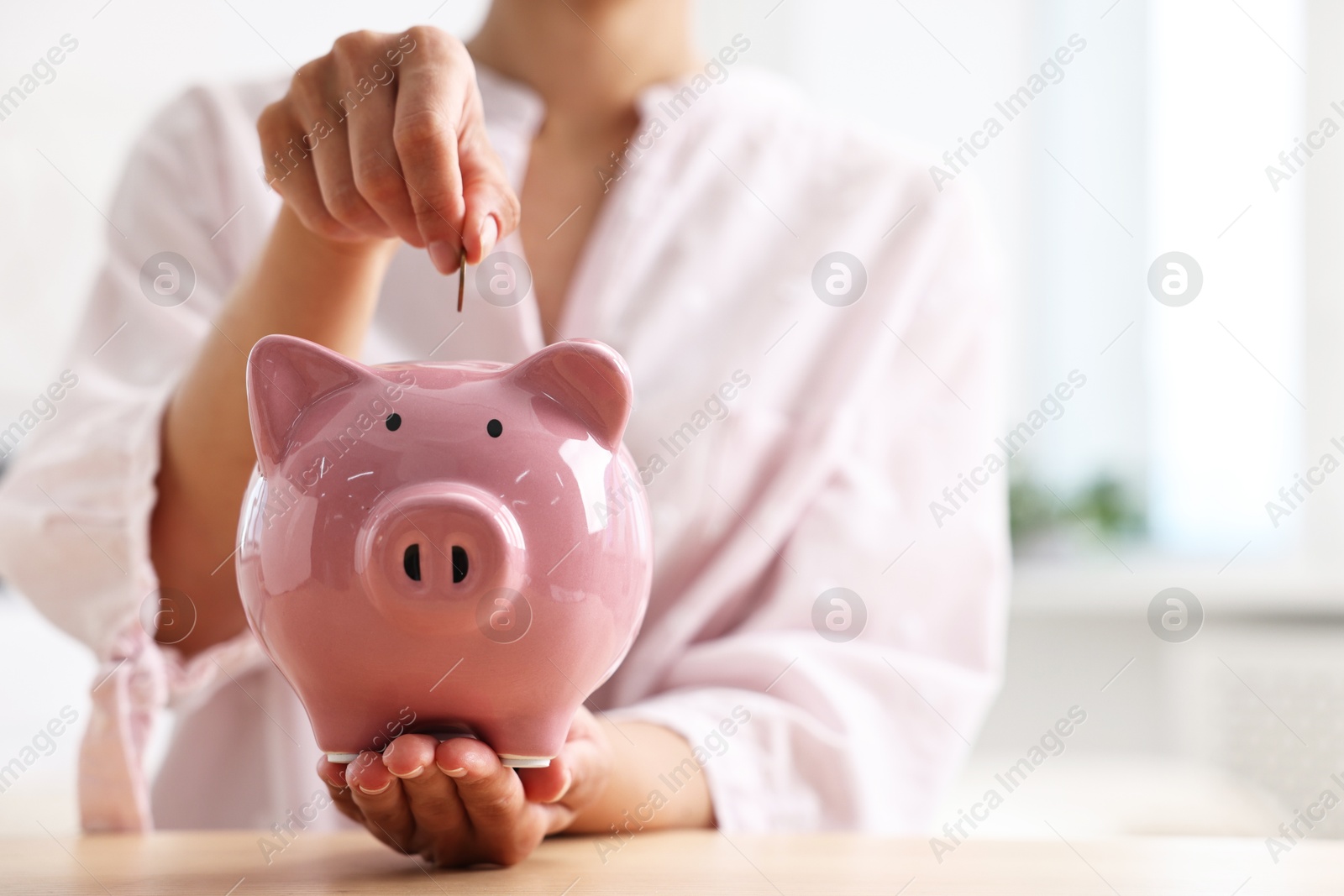 Photo of Woman putting coin into piggy bank at table indoors, closeup. Space for text