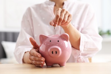 Woman putting coin into piggy bank at table indoors, closeup