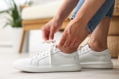 Photo of Woman tying shoelace of white sneaker indoors, closeup