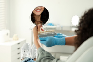 Patient looking in mirror while doctor holding teeth color chart in clinic, closeup. Dental veneers