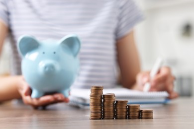 Photo of Woman with piggy bank taking notes at wooden table indoors, focus on stacks of coins