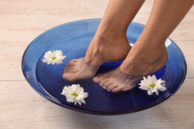 Photo of Woman soaking her feet in bowl with water and flowers on floor, closeup. Body care