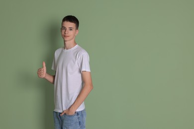 Teenage boy wearing white t-shirt and showing thumbs up on green background, space for text