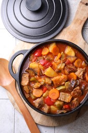 Delicious stew with vegetables in pot and spoon on light textured table, flat lay