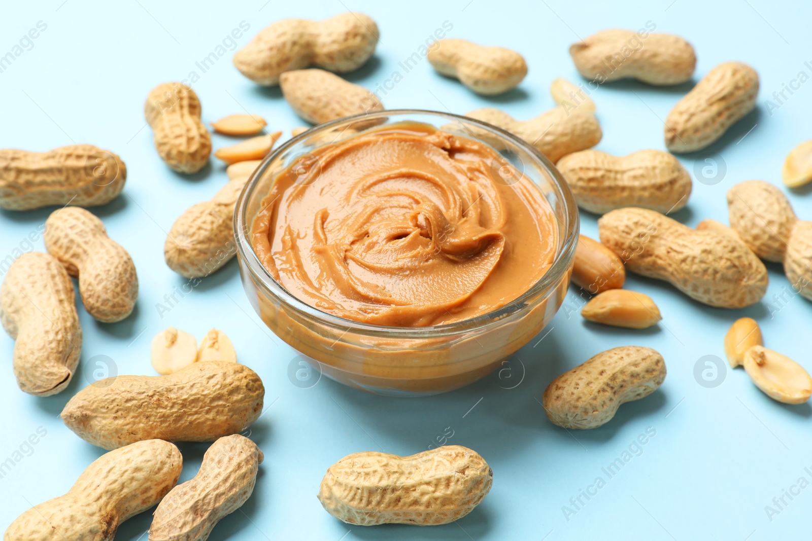 Photo of Tasty peanut butter in bowl and groundnuts on light blue background, closeup