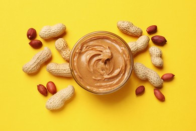 Tasty peanut butter in bowl and groundnuts on yellow table, flat lay