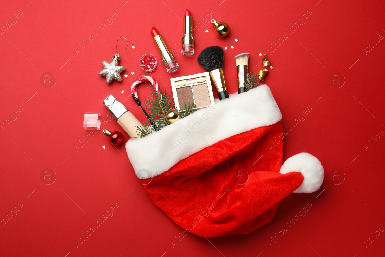 Photo of Santa hat with makeup products and Christmas decor on red background, top view