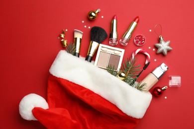 Photo of Santa hat with makeup products and Christmas decor on red background, top view