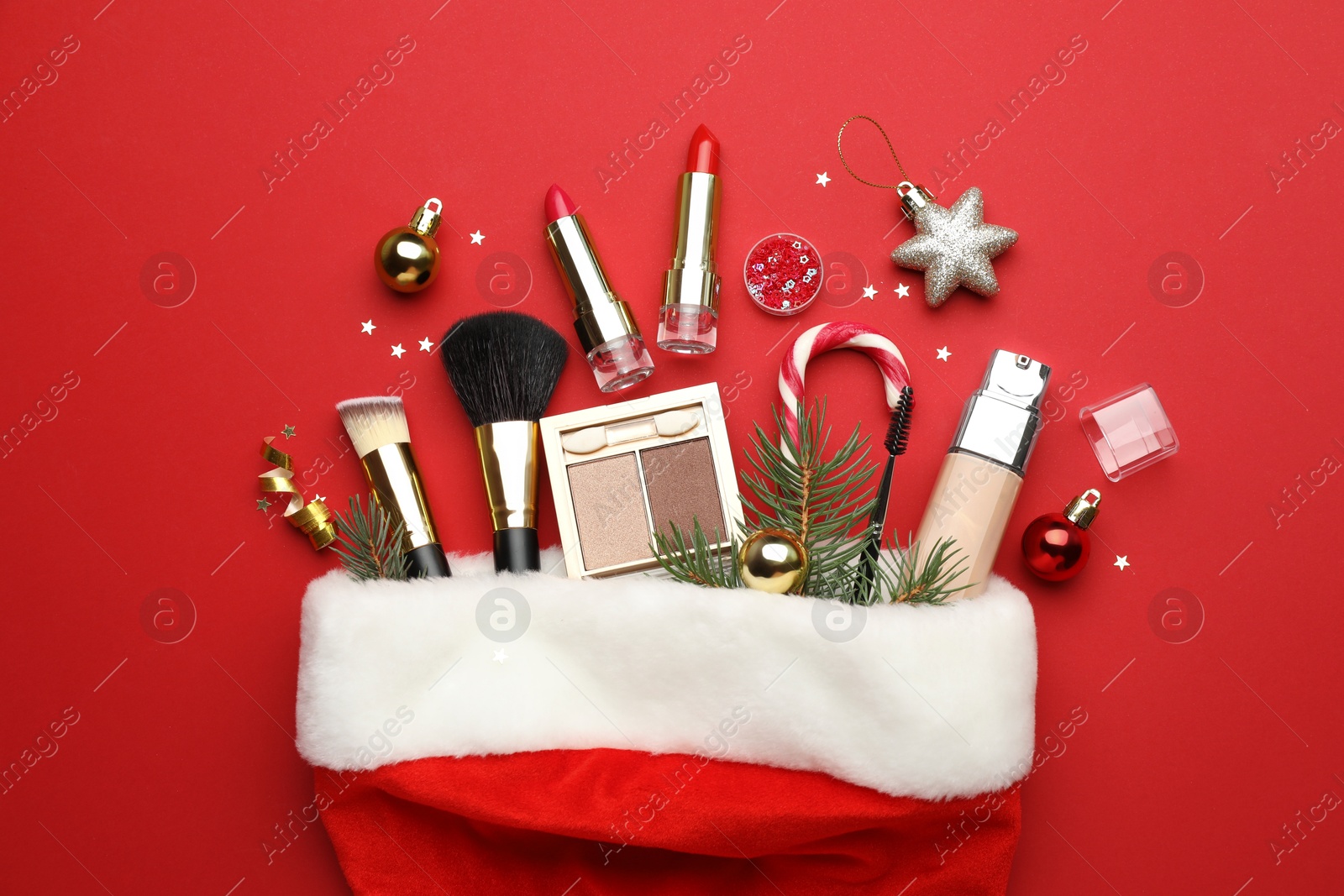 Photo of Santa hat with makeup products and Christmas decor on red background, top view