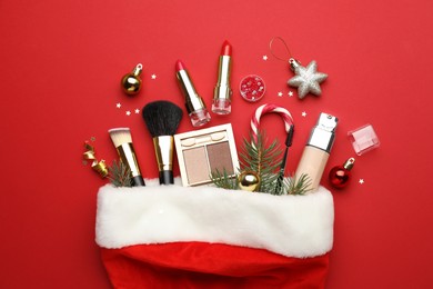 Santa hat with makeup products and Christmas decor on red background, top view