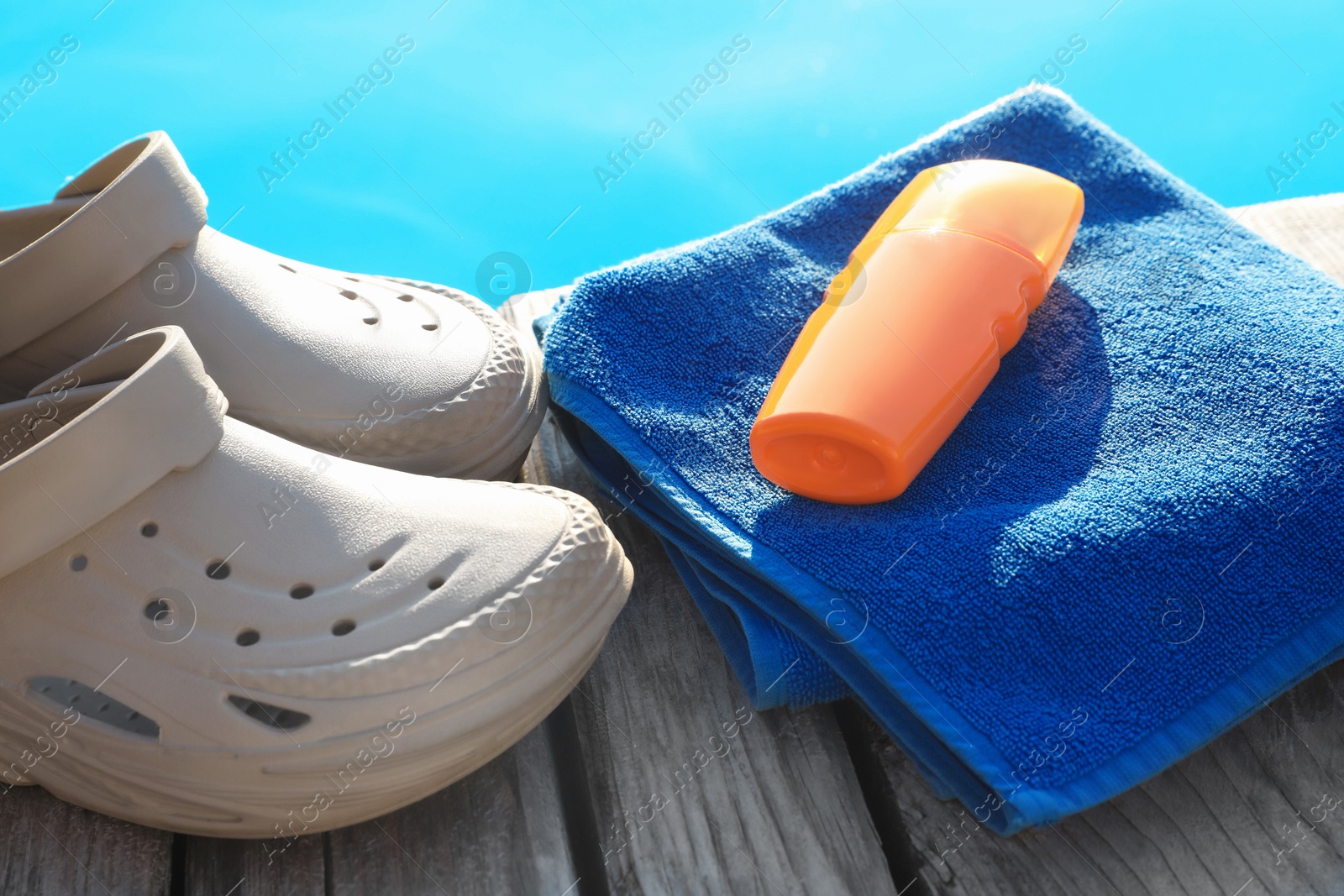 Photo of Stylish beach slippers, towel and sunscreen on wooden deck near outdoor swimming pool, closeup