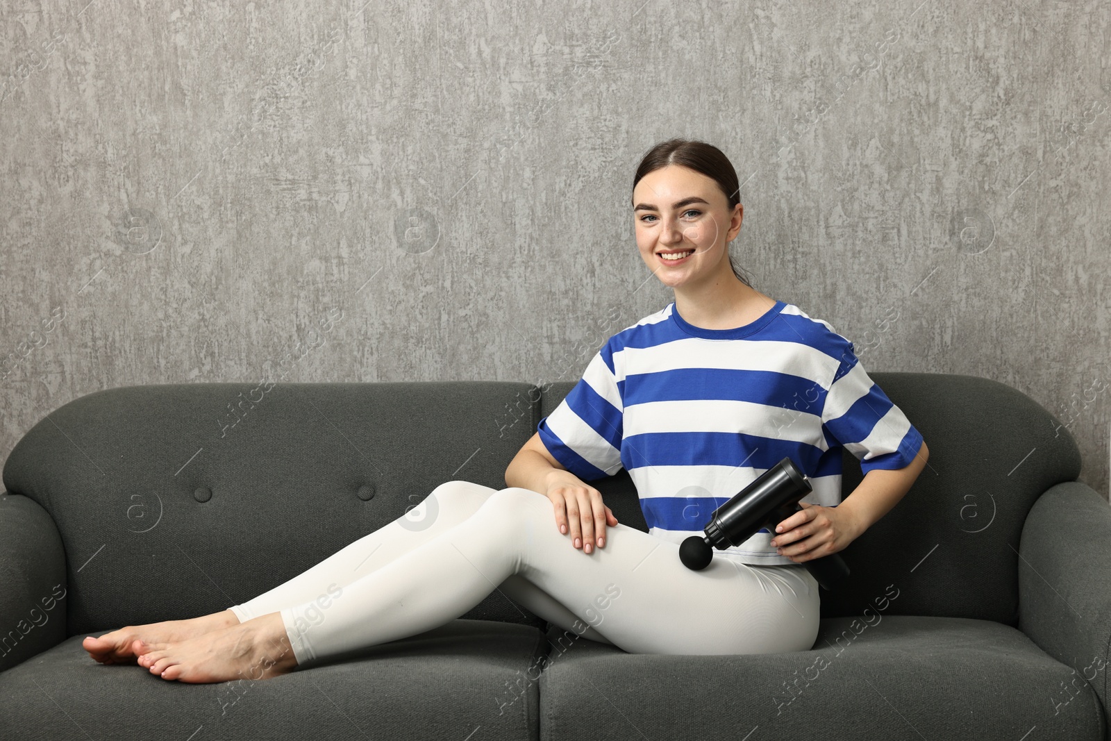 Photo of Woman using percussive massager to relieve hip muscles on sofa indoors