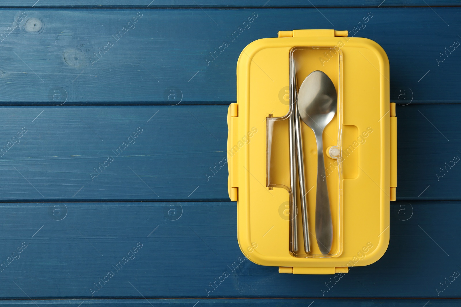 Photo of Yellow lunch box with cutlery on blue wooden table, top view. Space for text