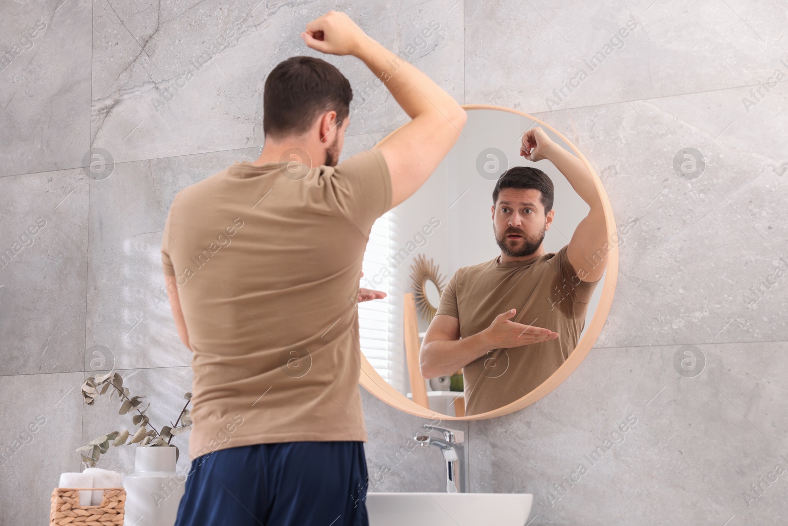 Photo of Emotional man in t-shirt before using deodorant near mirror at home, back view