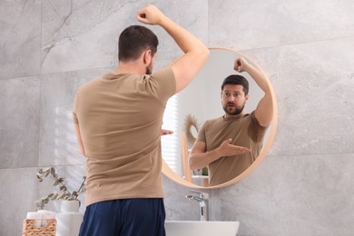 Photo of Emotional man in t-shirt before using deodorant near mirror at home, back view