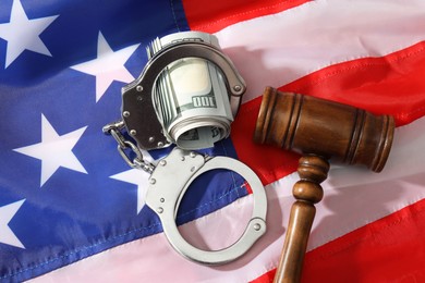 Handcuffs with dollar banknotes and wooden gavel on American flag, flat lay