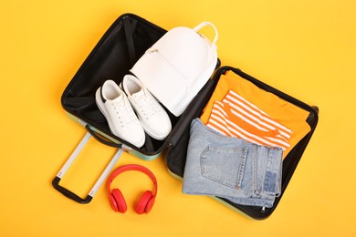 Photo of Open suitcase with backpack, sneakers, headphones and clothes on yellow background, above view