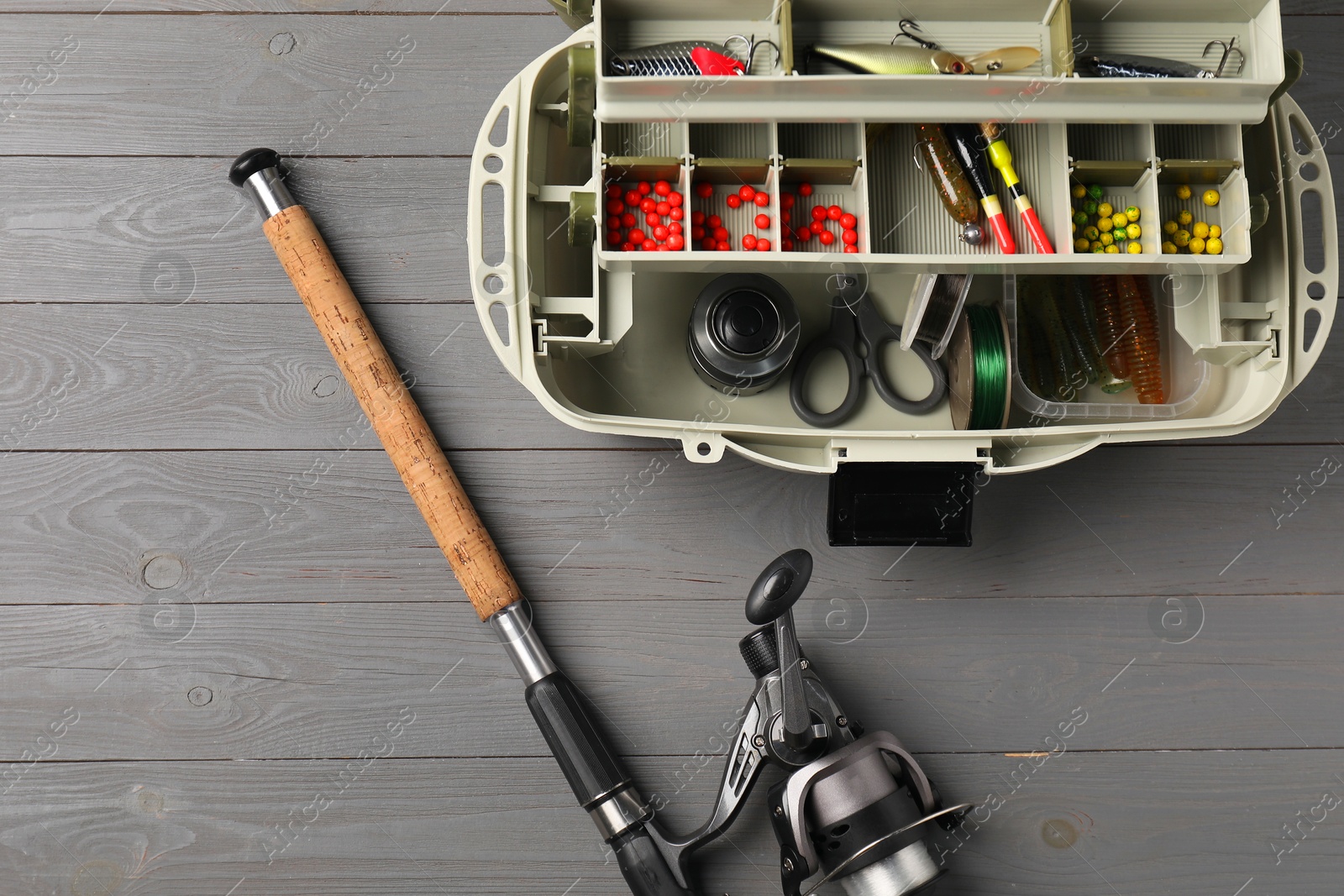 Photo of Different fishing tackles in box and rod with spinning reel on grey wooden table, top view. Space for text