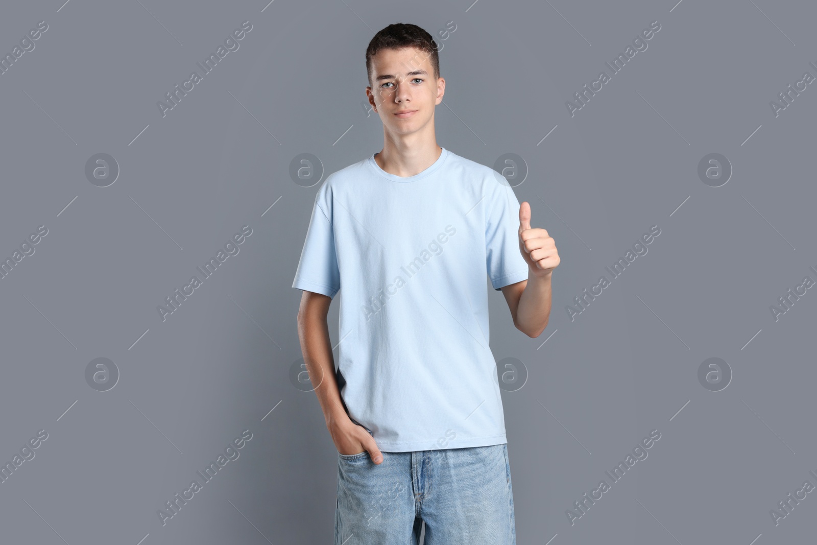 Photo of Teenage boy wearing light blue t-shirt and showing thumbs up on grey background