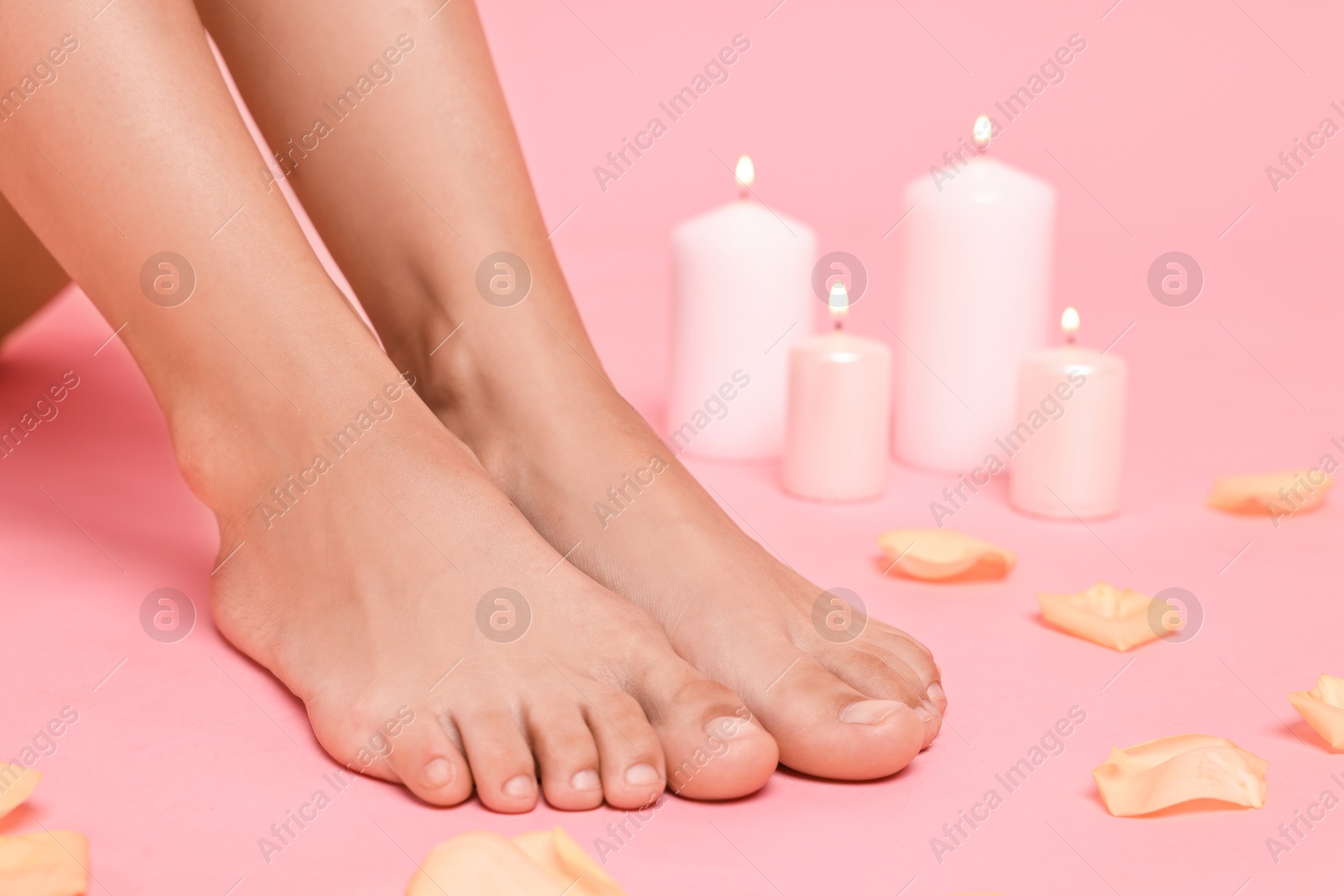 Photo of Woman with smooth feet, burning candles and petals on pink background, closeup