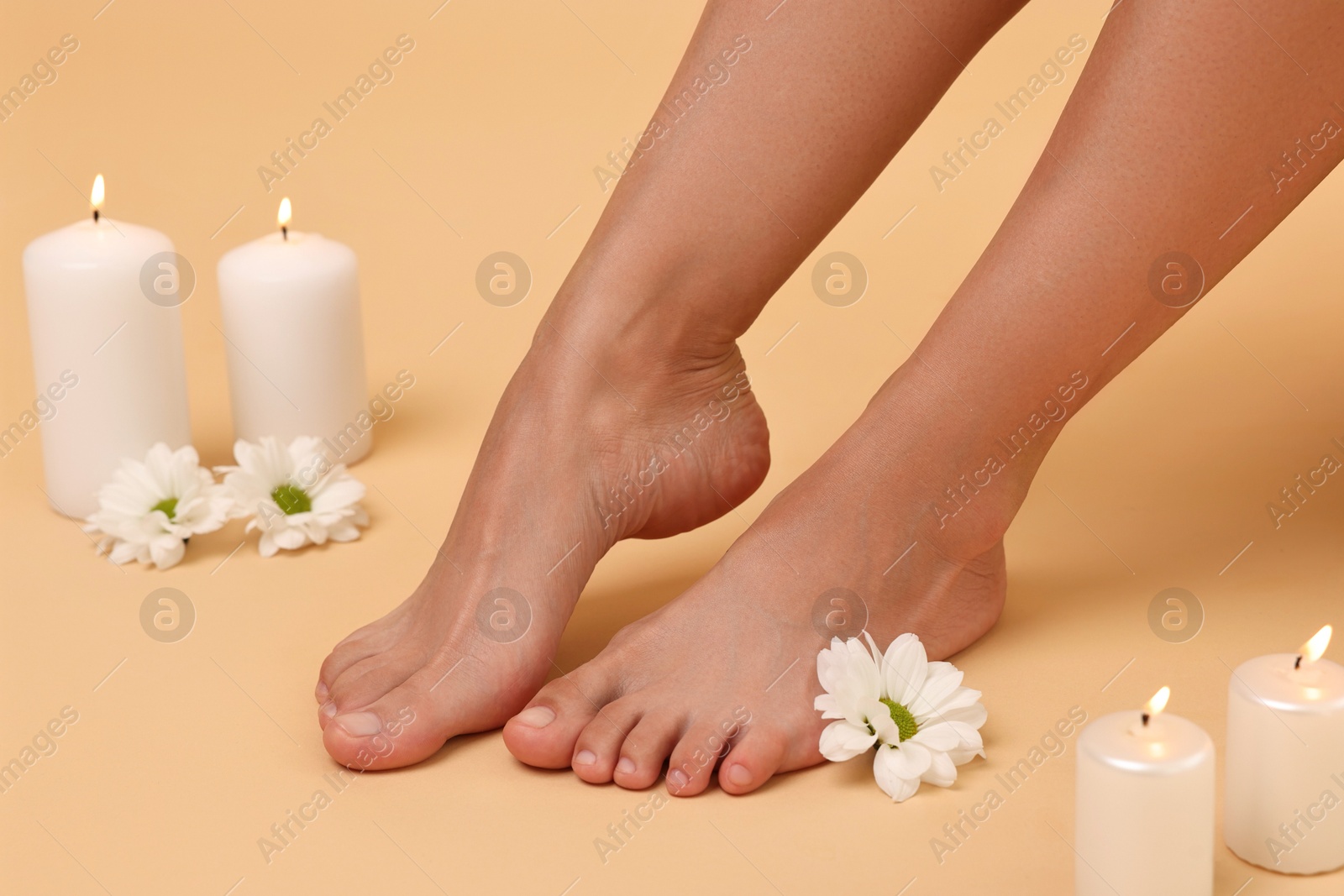 Photo of Woman with smooth feet, burning candles and chrysanthemum flowers on beige background, closeup