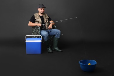 Fisherman with rod and cooler box fishing from bowl on chair against black background