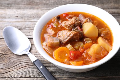 Delicious stew with vegetables in bowl and spoon on wooden table, closeup