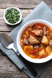 Delicious stew with vegetables in bowl, parsley and spoon on wooden table, top view