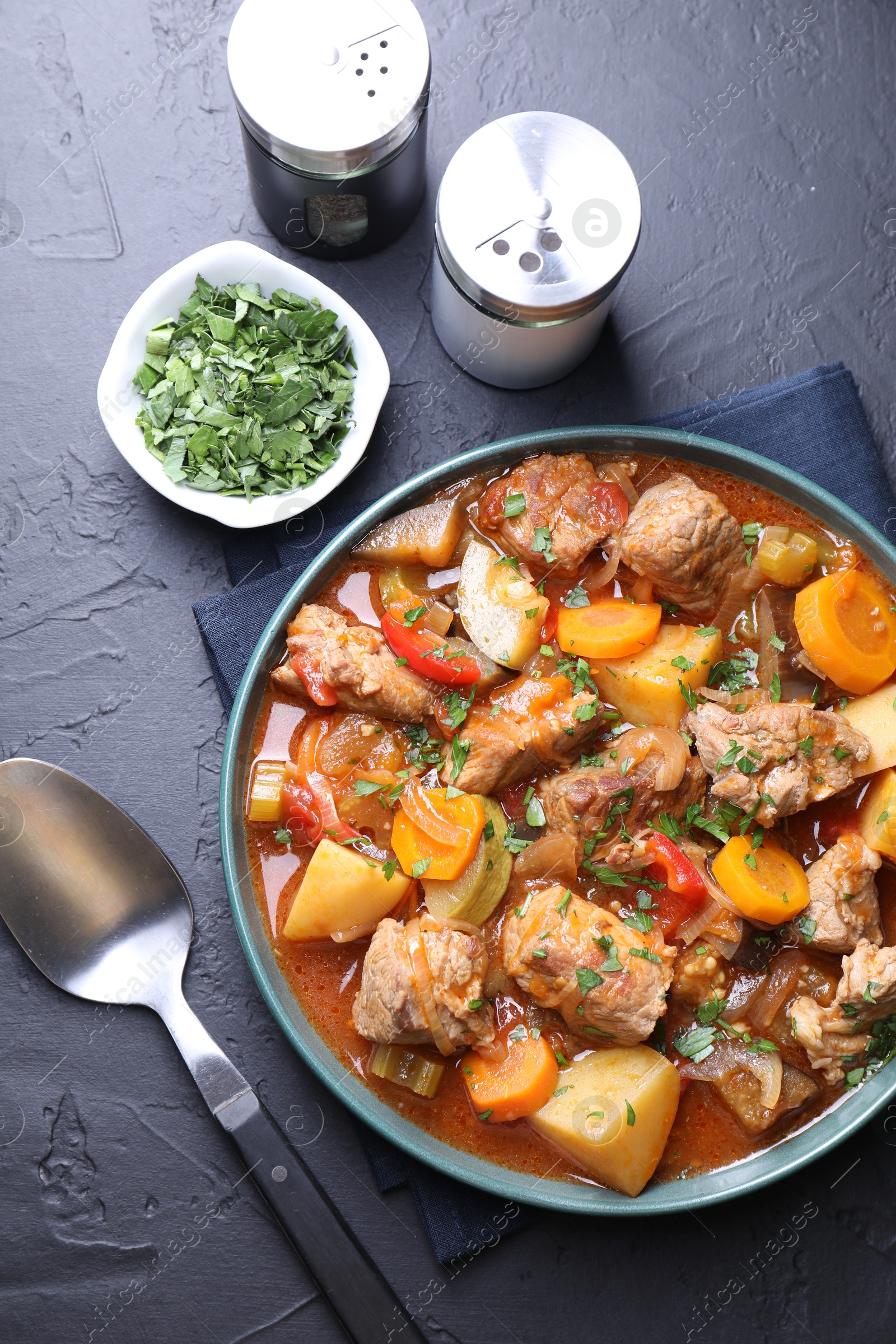 Photo of Delicious stew with vegetables, parsley and spoon on black table, top view