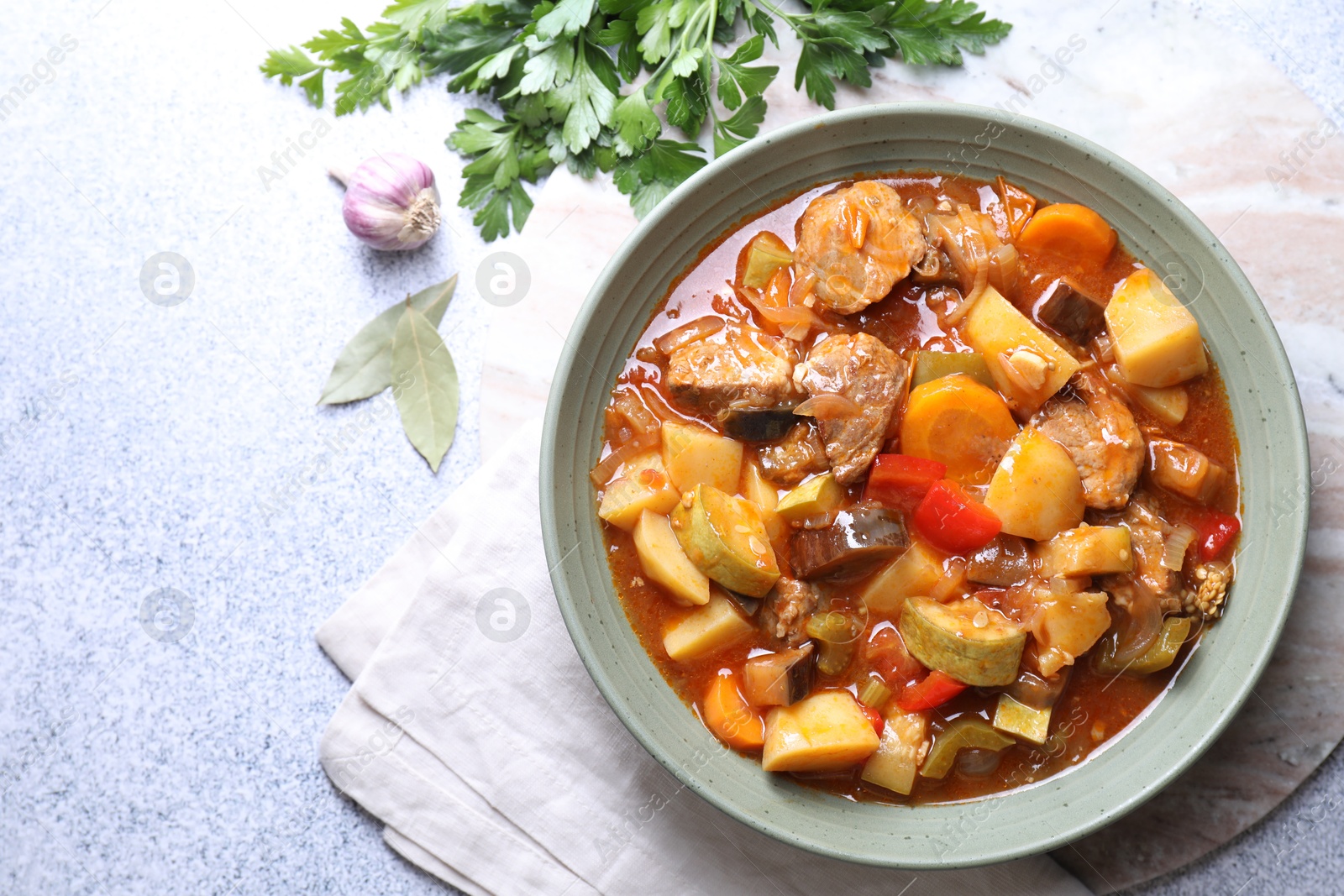 Photo of Delicious stew with vegetables in bowl, parsley, garlic and bay leaves on light grey table, top view