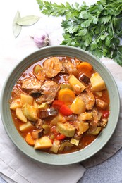 Photo of Delicious stew with vegetables in bowl, parsley, garlic and bay leaves on light grey table, top view