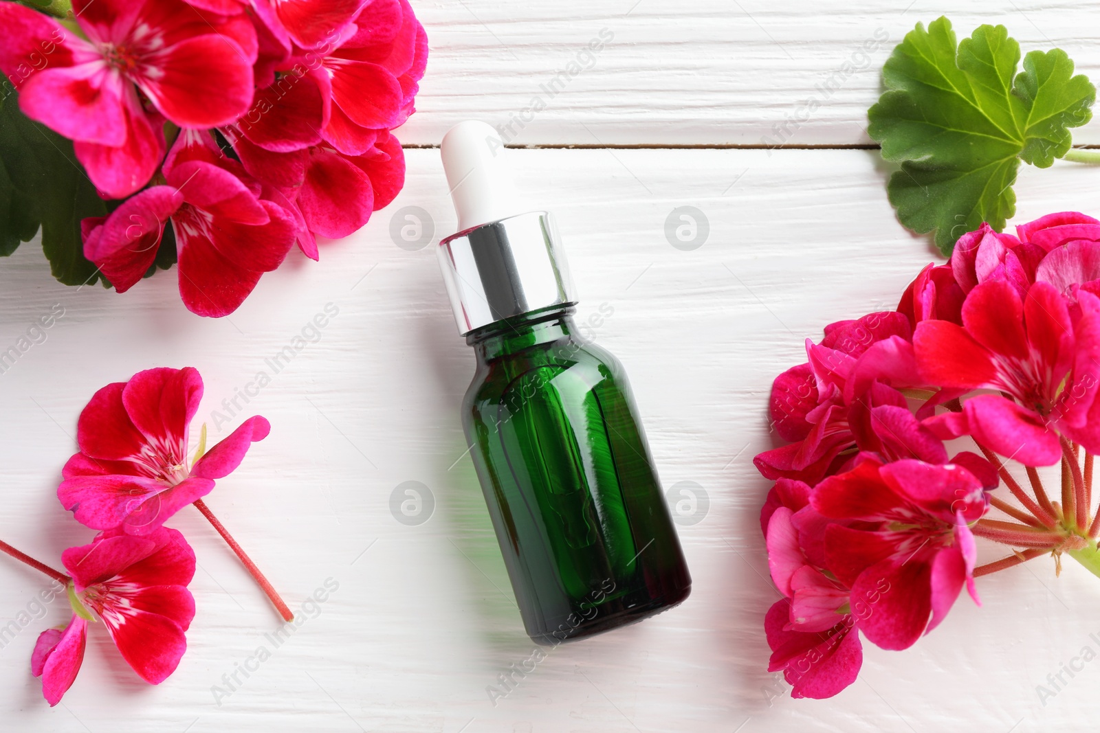 Photo of Bottle of geranium essential oil and beautiful flowers on white wooden table, flat lay