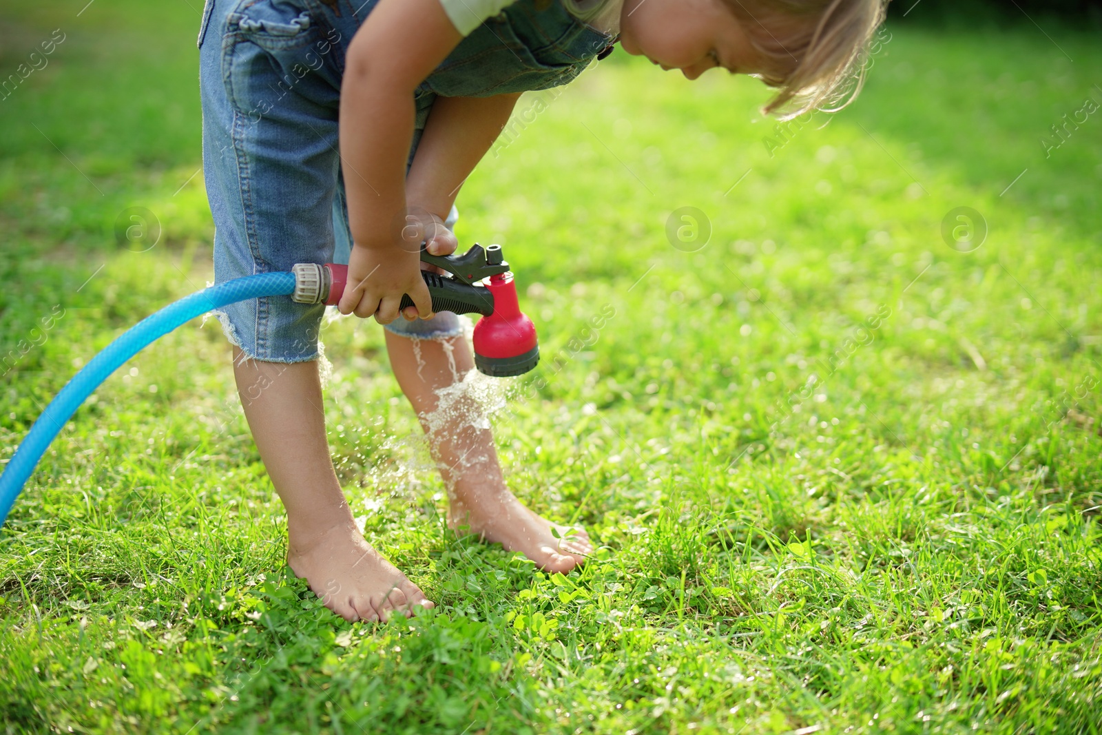 Photo of Little girl watering green grass on lawn in backyard, closeup