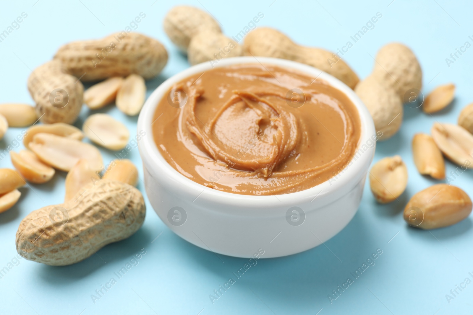 Photo of Tasty peanut butter in bowl and groundnuts on light blue background, closeup