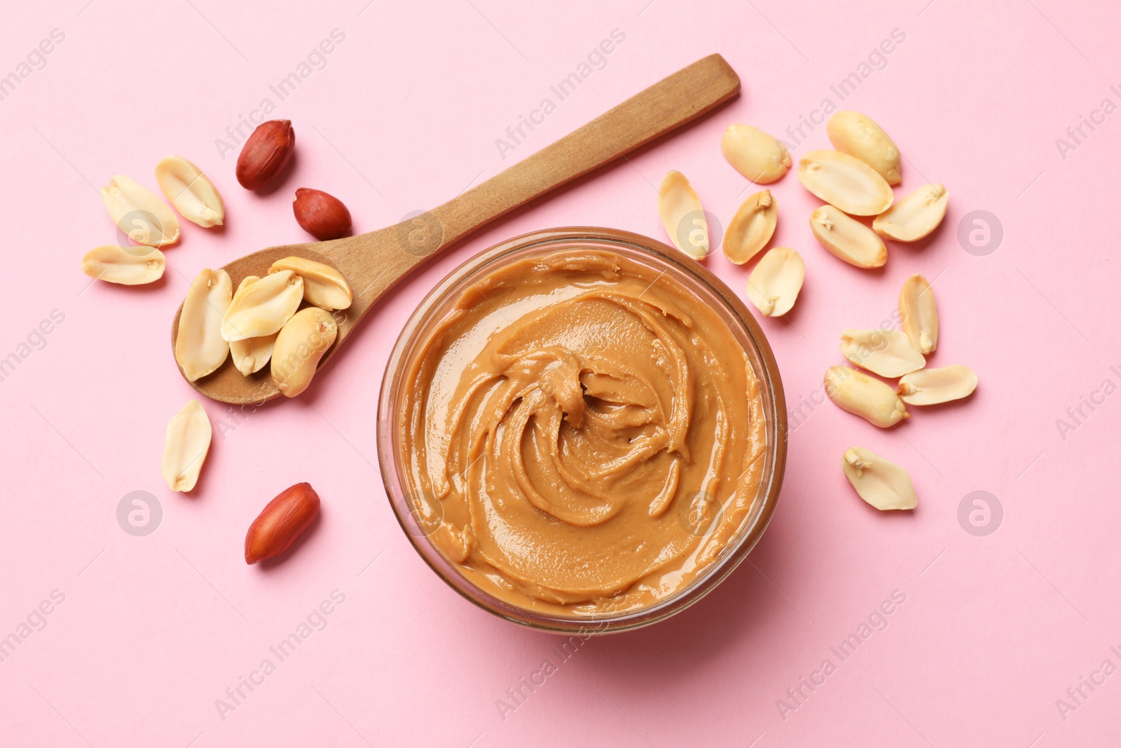 Photo of Tasty peanut butter in bowl and spoon with groundnuts on pink table, flat lay