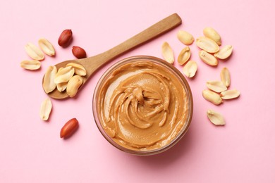 Photo of Tasty peanut butter in bowl and spoon with groundnuts on pink table, flat lay