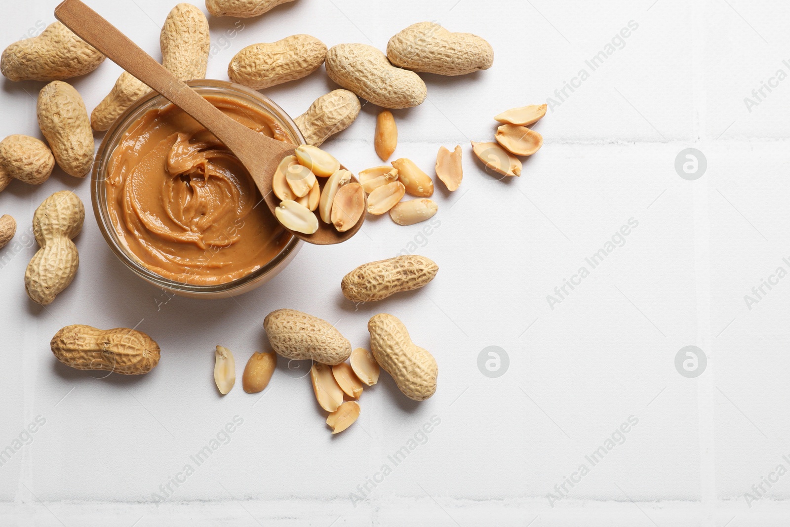 Photo of Tasty peanut butter in bowl and groundnuts on white tiled table, flat lay. Space for text