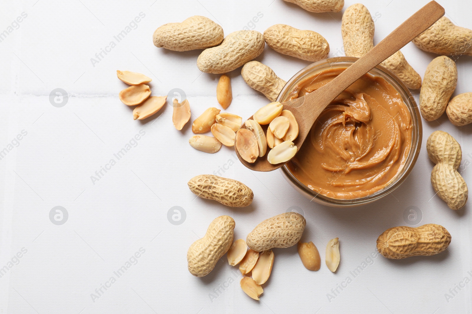 Photo of Tasty peanut butter in bowl and groundnuts on white tiled table, flat lay. Space for text