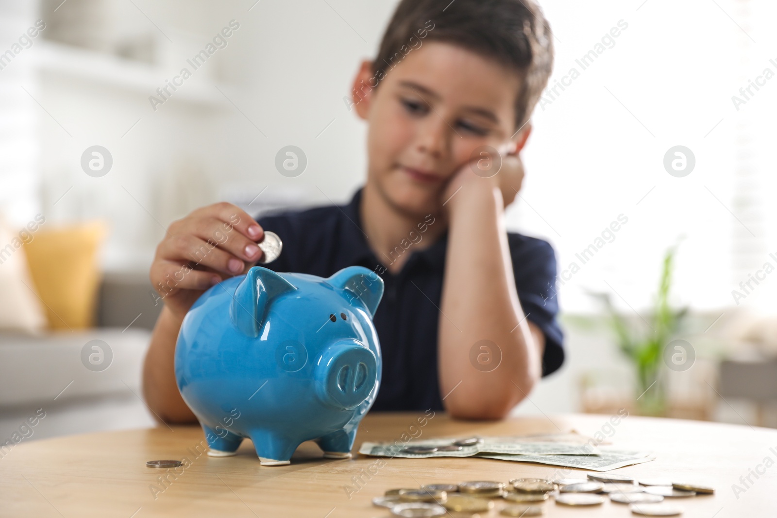 Photo of Pocket money. Cute boy putting coin into piggy bank at wooden table indoors, selective focus